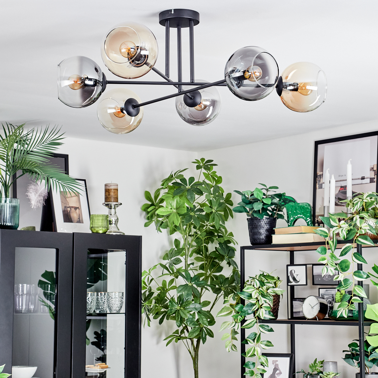 Ein modernes Wohnzimmer mit schwarzem Kronleuchter, Pflanzen, gerahmten Bildern und einem schwarzen Schrank. Die Decke ist weiß.