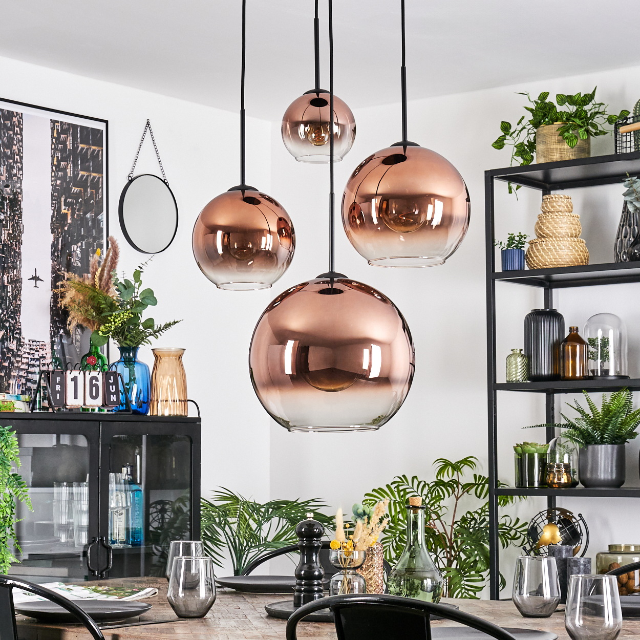 Esszimmer mit kupferfarbenen Pendelleuchten. Tisch mit Gläsern. Pflanzen und Regale mit Dekor im Hintergrund.