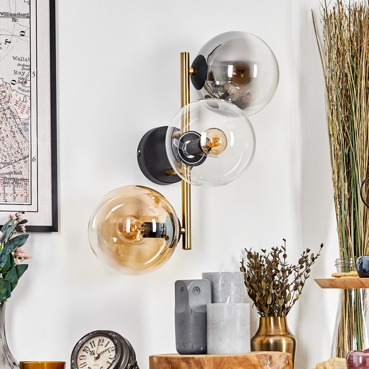 Eine Wandlampe mit drei Glaskugeln, einem goldenen Rahmen und einem schwarzen Sockel ist an einer weißen Wand befestigt. Im Vordergrund befinden sich verschiedene Dekorationsartikel.