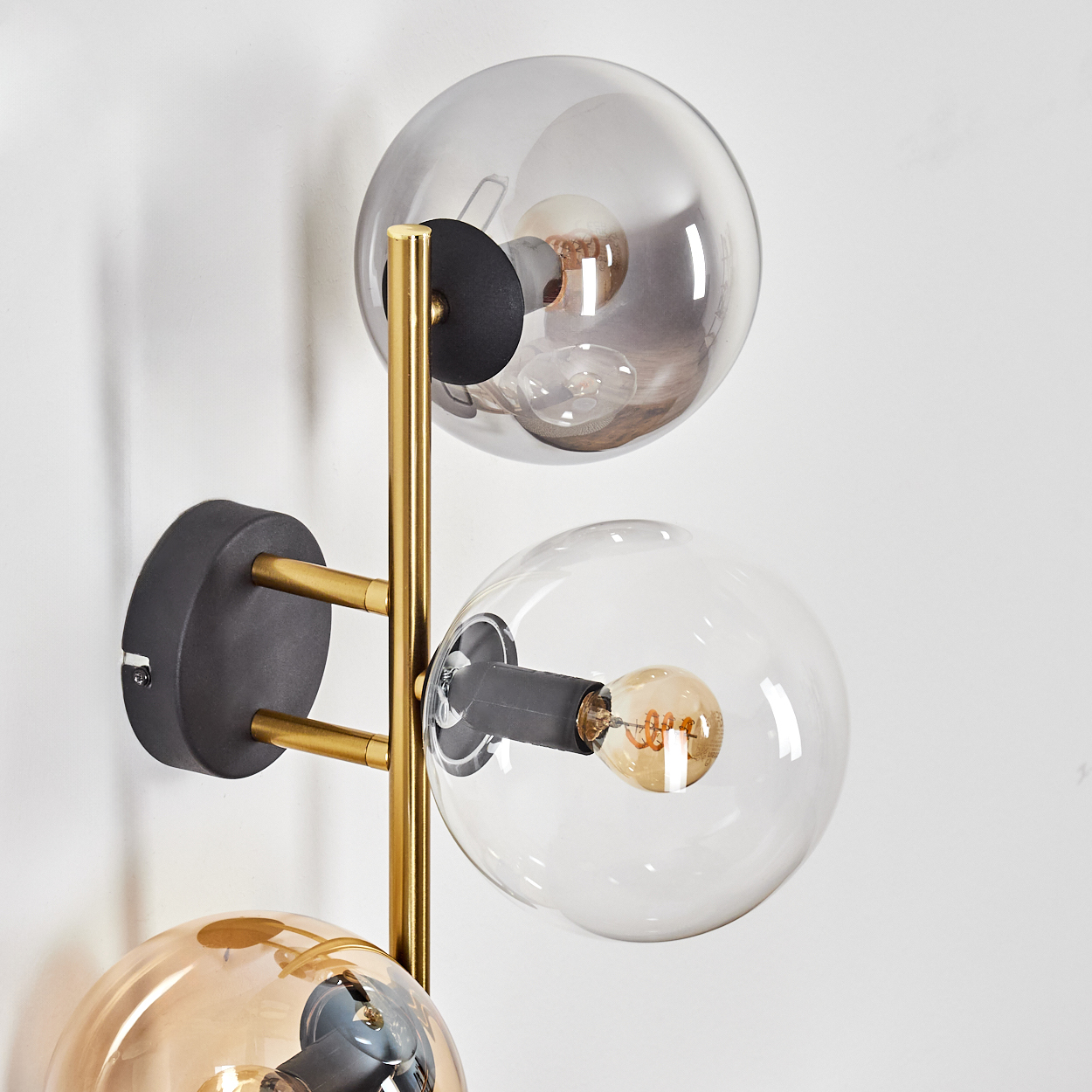 Eine moderne Wandlampe mit drei kugelförmigen Glasschirmen. Goldene Metallstäbe und schwarzer Sockel, Glühbirnen im Inneren. Weißer Wandhintergrund.