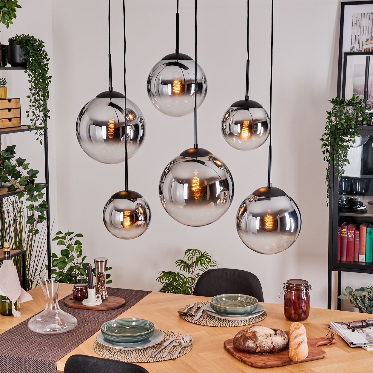 Esszimmer mit Holztisch. Sechs Pendelleuchten hängen darüber. Teller, Brot und Pflanzen schmücken die Szene.