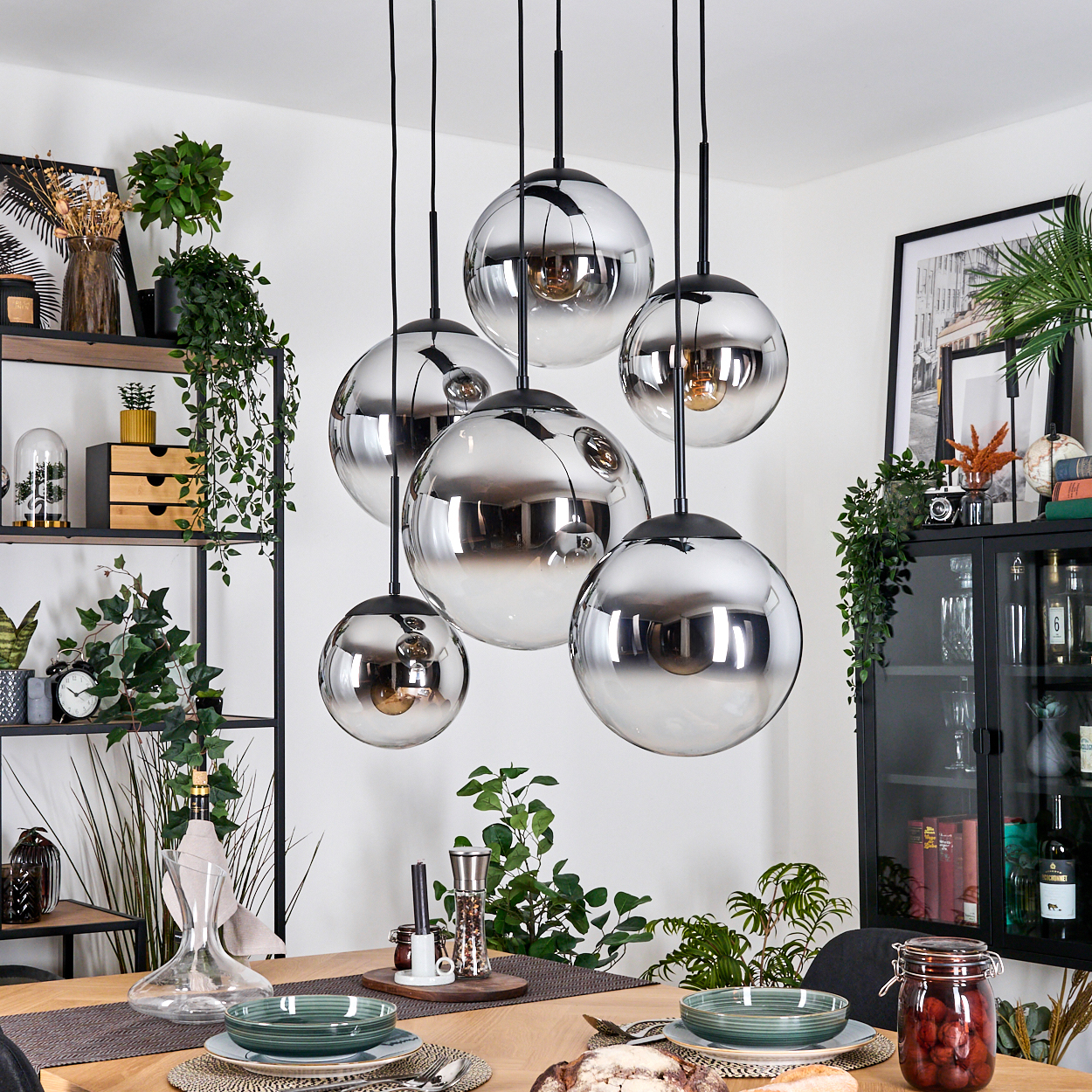 Esszimmer mit hängenden Kugelleuchten. Tisch mit Tellern, Pflanzen und einem schwarzen Schrank mit Dekor.