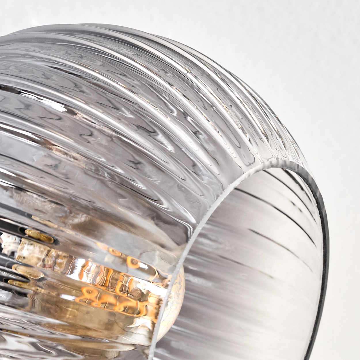 Nahaufnahme eines Lampenschirms aus Glas mit gerippter Textur und einer Glühbirne im Inneren. Das Glas ist transparent, das Licht warm.