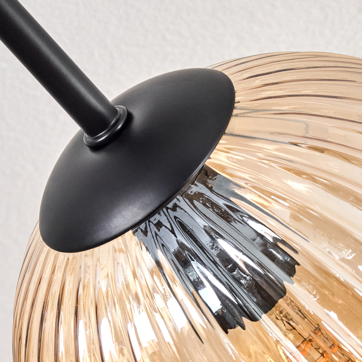 Nahaufnahme einer Lampe. Mit schwarzem Arm und Schirm, mit geripptem, transparentem, bernsteinfarbenem Glaskörper. Hintergrund ist eine weiße Wand.