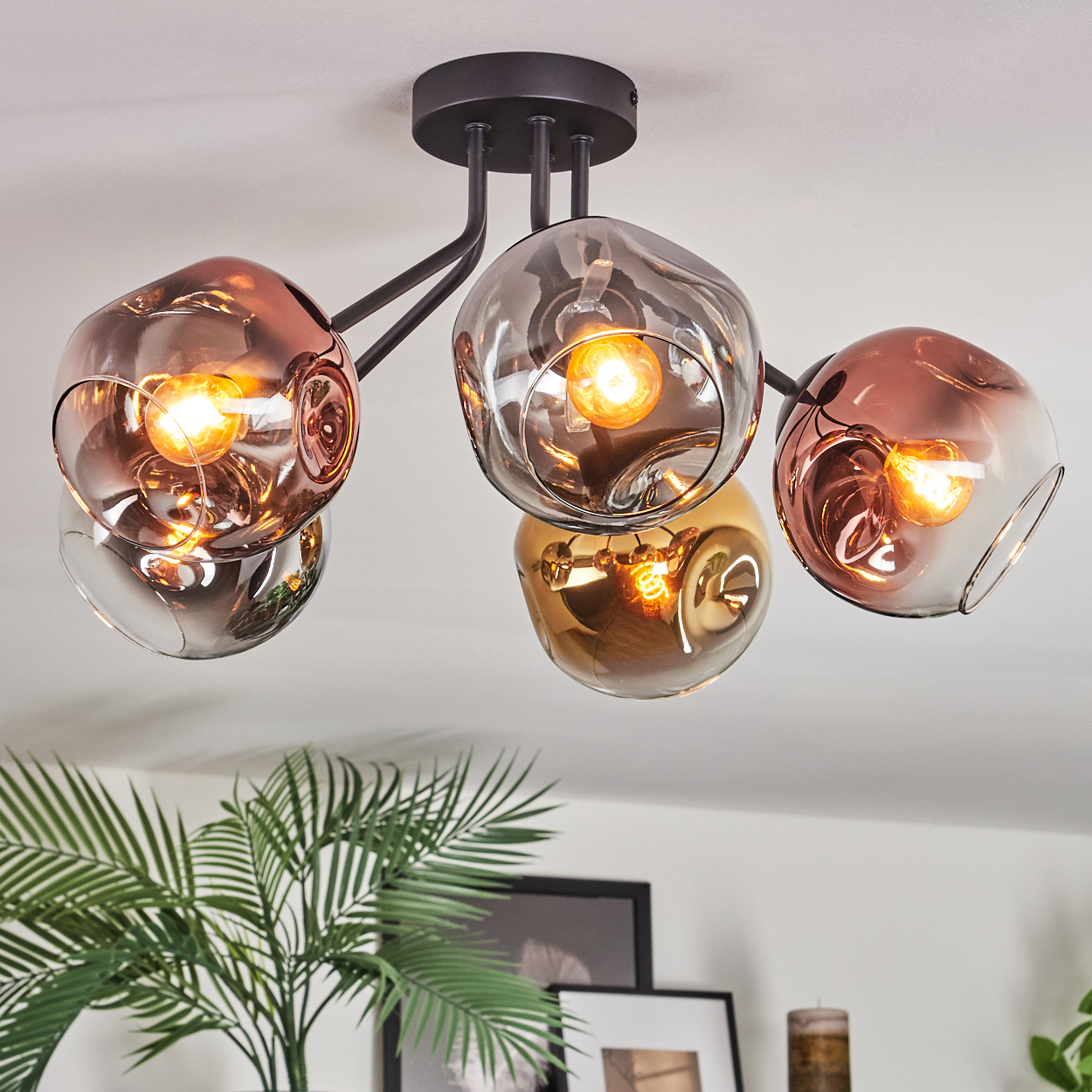 Eine moderne Deckenleuchte mit fünf Glasschirmen in Kupfer und Grau. Im Hintergrund sind eine grüne Pflanze und gerahmte Bilder zu sehen.