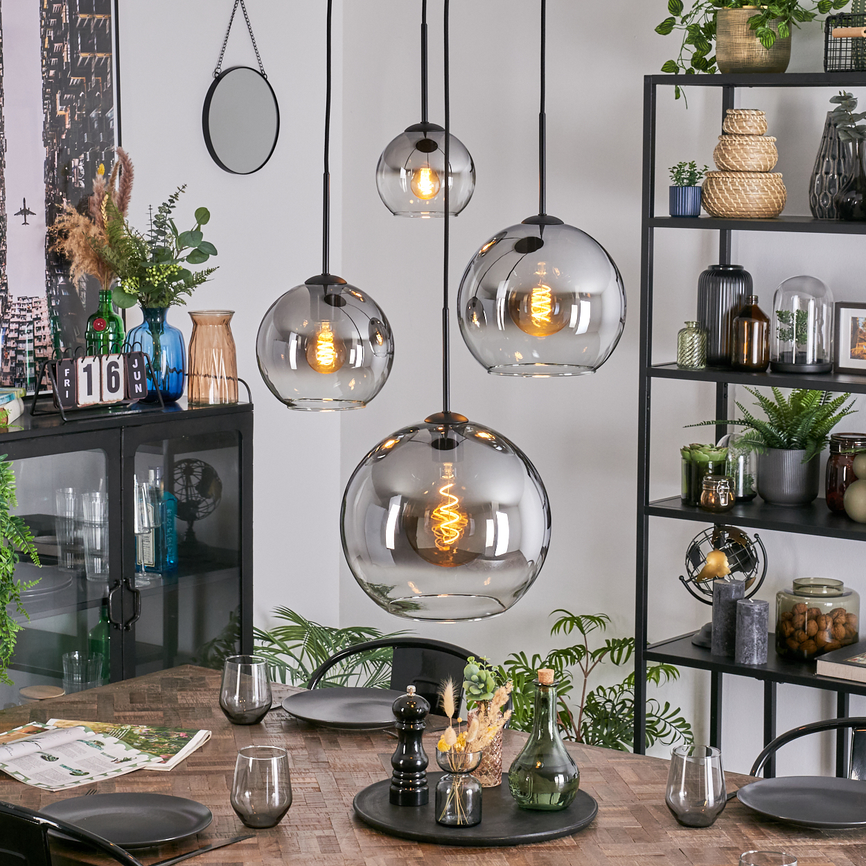 Esszimmer mit hängenden Kugelleuchten. Tisch mit Tellern, Gläsern und Dekorationen. Regale mit Pflanzen und Dekoration.