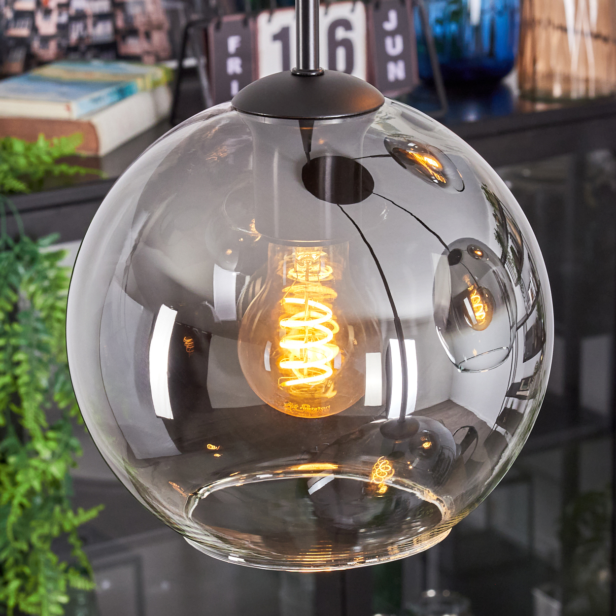 Eine moderne Glaskugel-Pendelleuchte mit einer Wendelglühbirne im Inneren. Die Kugel reflektiert die Umgebung. Im Hintergrund ist ein Schreibtischkalender zu sehen.