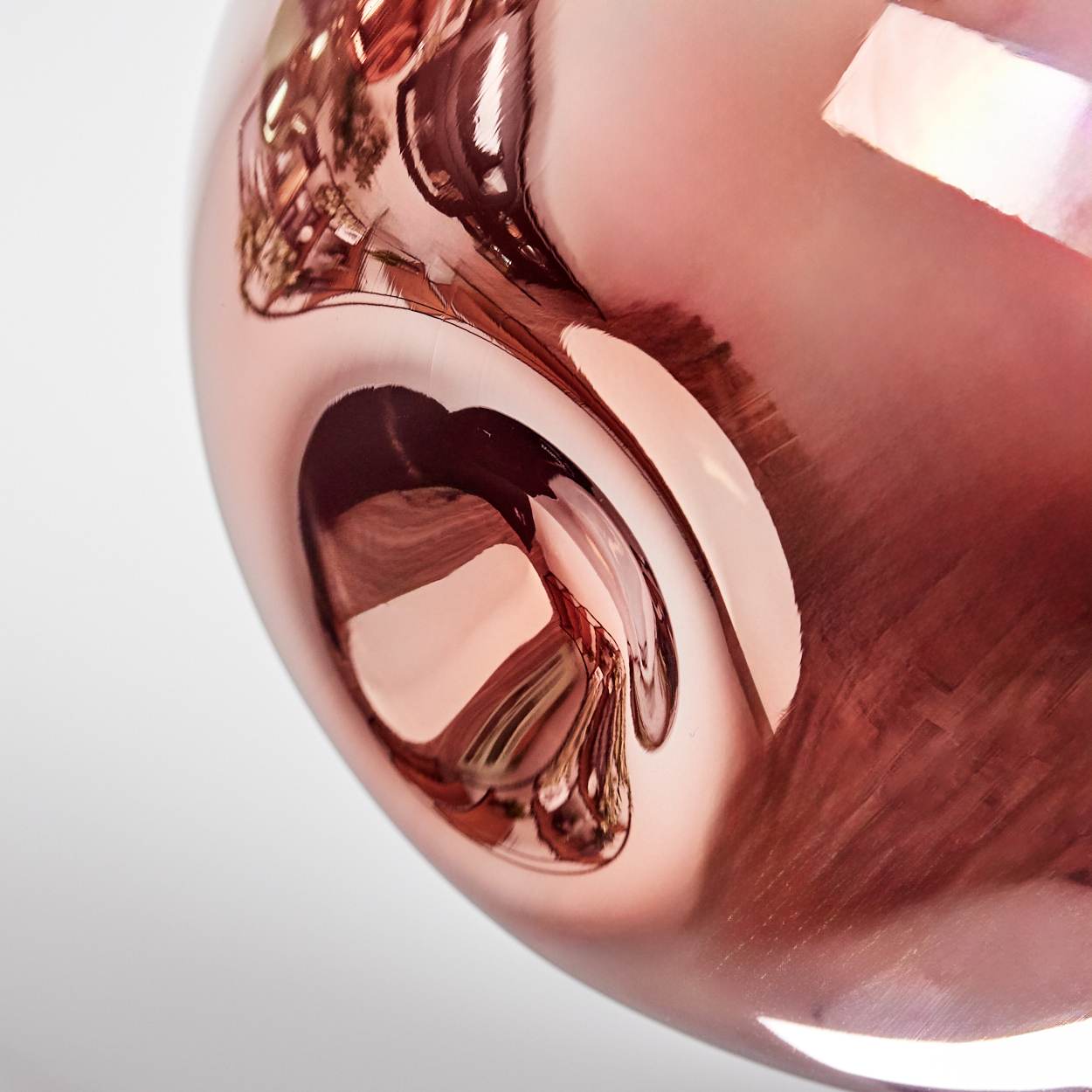 Nahaufnahme einer reflektierenden, roségoldenen Metalloberfläche. Reflexionen verzerren das Bild, zeigen Licht und Formen. Weißer Hintergrund.
