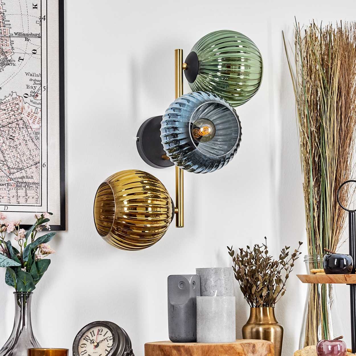 Eine Wandlampe mit drei Glaskugeln in Grün, Blau und Gold. Eine Karte, eine Vase mit Blumen, Kerzen und dekorative Gegenstände sind ebenfalls vorhanden.
