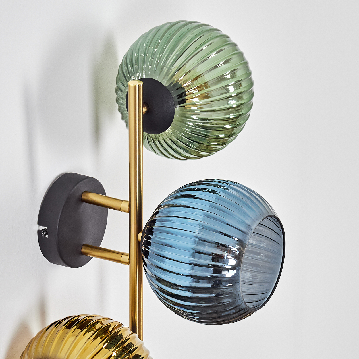 Eine moderne Wandlampe mit drei kugelförmigen Glasschirmen in Grün, Blau und Gold. Die Lampe hat ein goldenes Gestell und einen schwarzen Sockel.