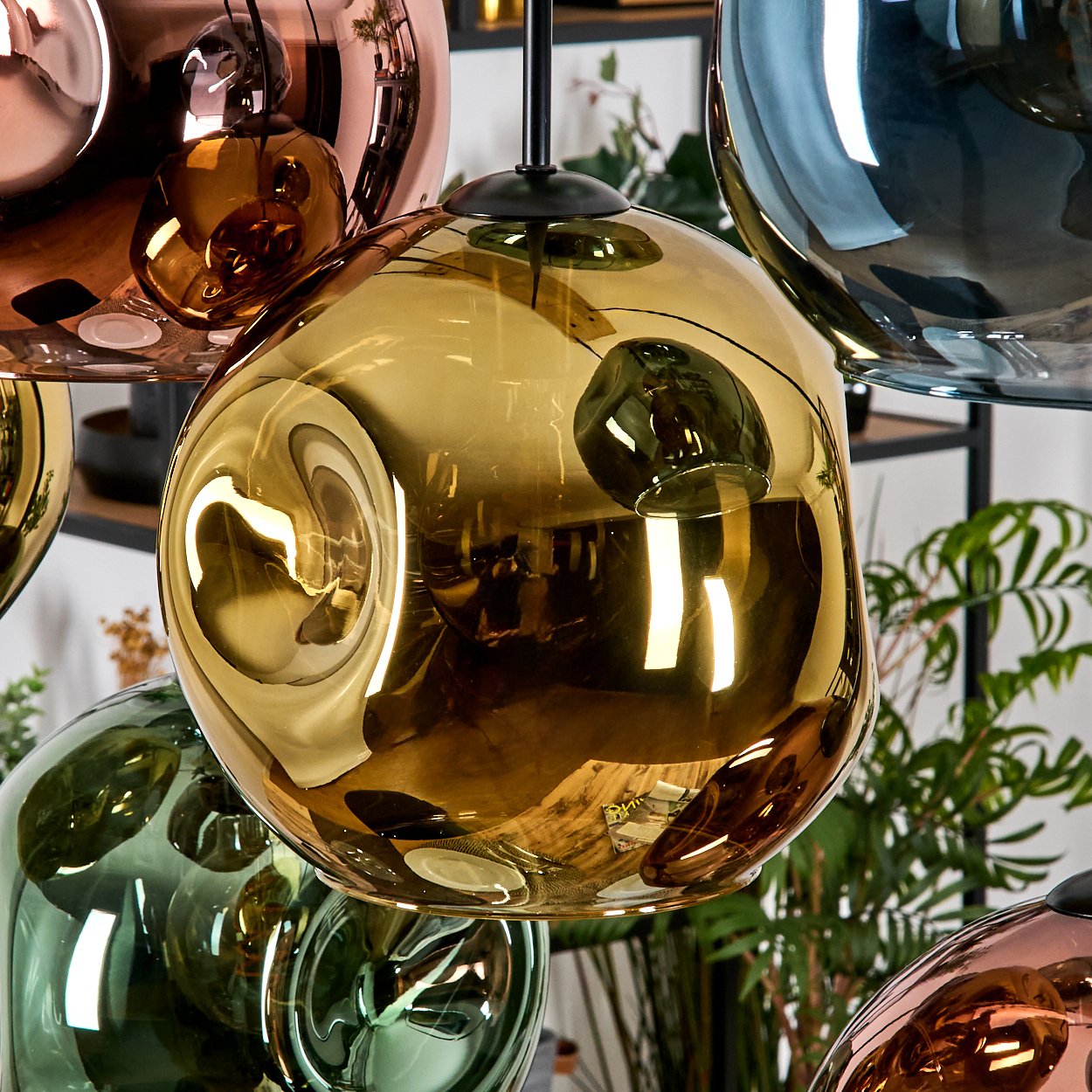 Nahaufnahme von Pendelleuchten mit reflektierenden Oberflächen in Gold, Kupfer, Grün und Blau. Grüne Pflanzen im Hintergrund.
