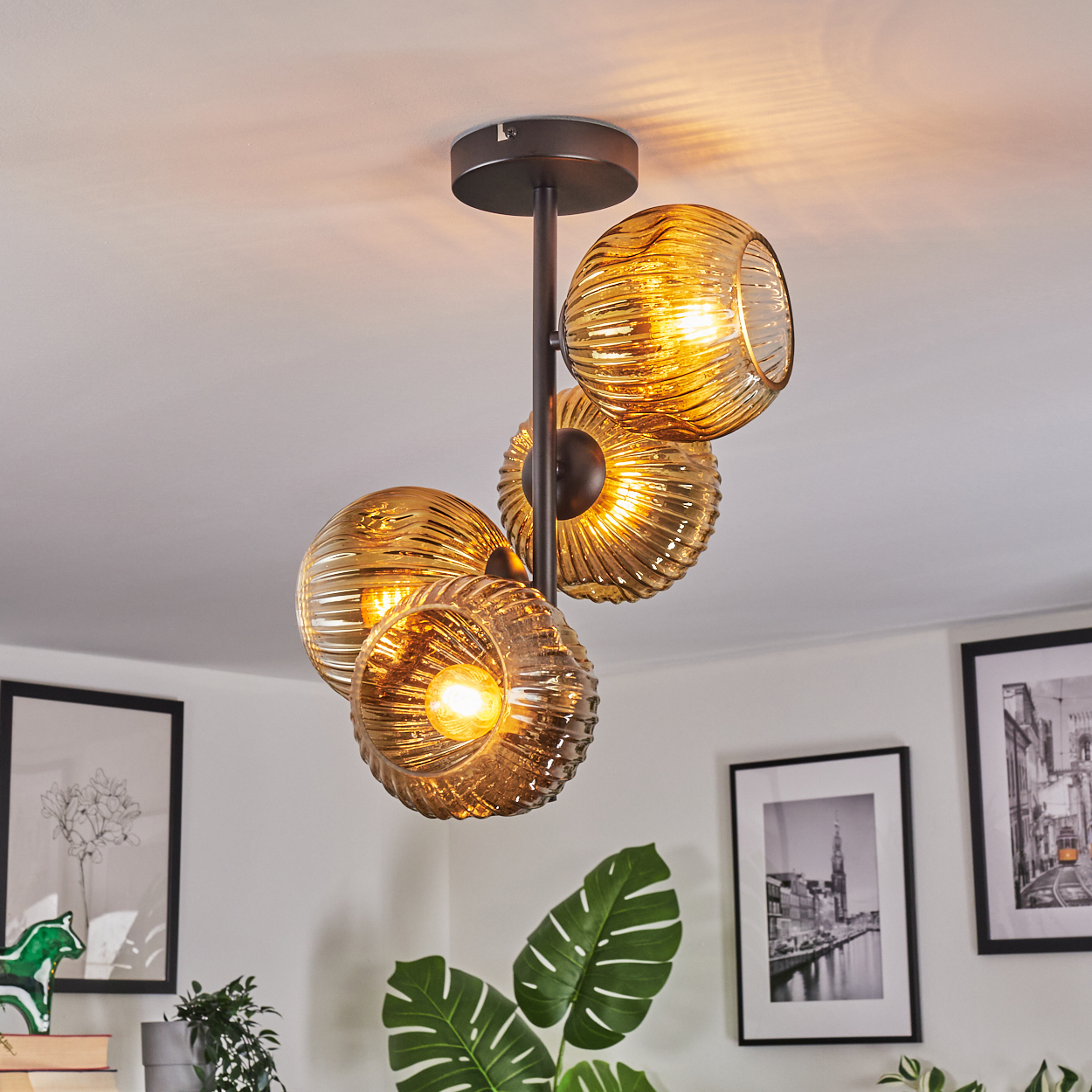 Eine moderne Deckenleuchte mit vier bernsteinfarbenen Glaskugeln. Das Licht ist an und beleuchtet den Raum. Im Hintergrund sind Bilder und Pflanzen zu sehen.