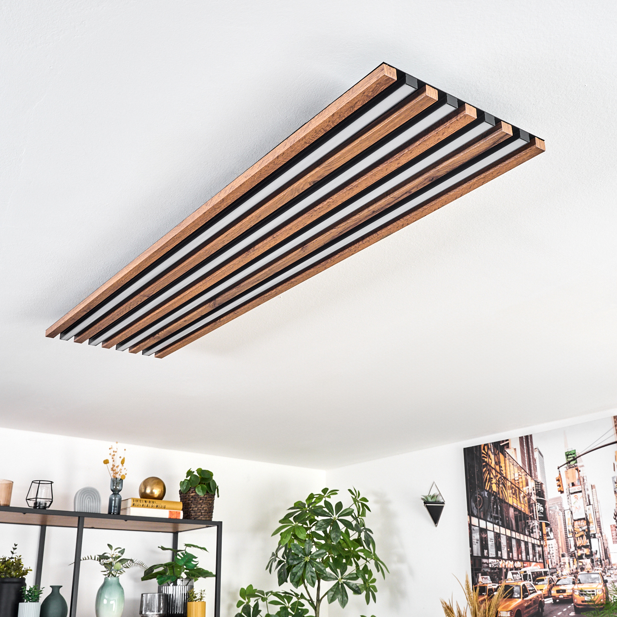 Eine moderne Deckenleuchte mit Holzlatten und weißen Lichtern. Darunter ein Raum mit Pflanzen, einem Regal und einem New York Poster.