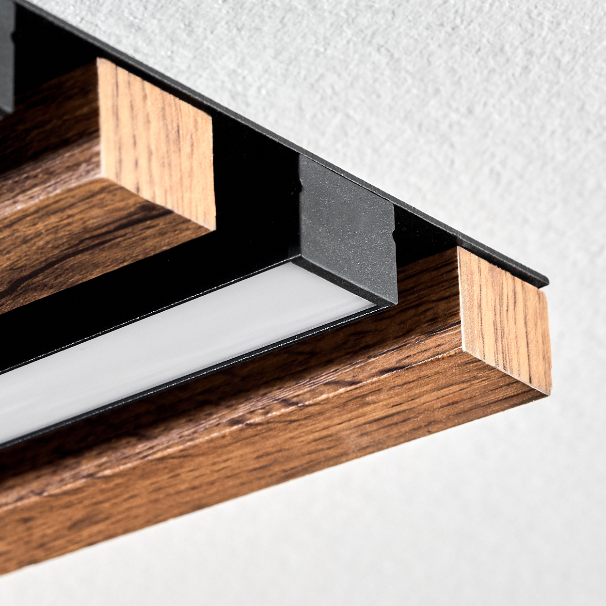 Nahaufnahme eines Holzbalkens mit schwarzen Metallakzenten und einem weißen Lichtstreifen. Der Hintergrund ist eine weiße Wand.
