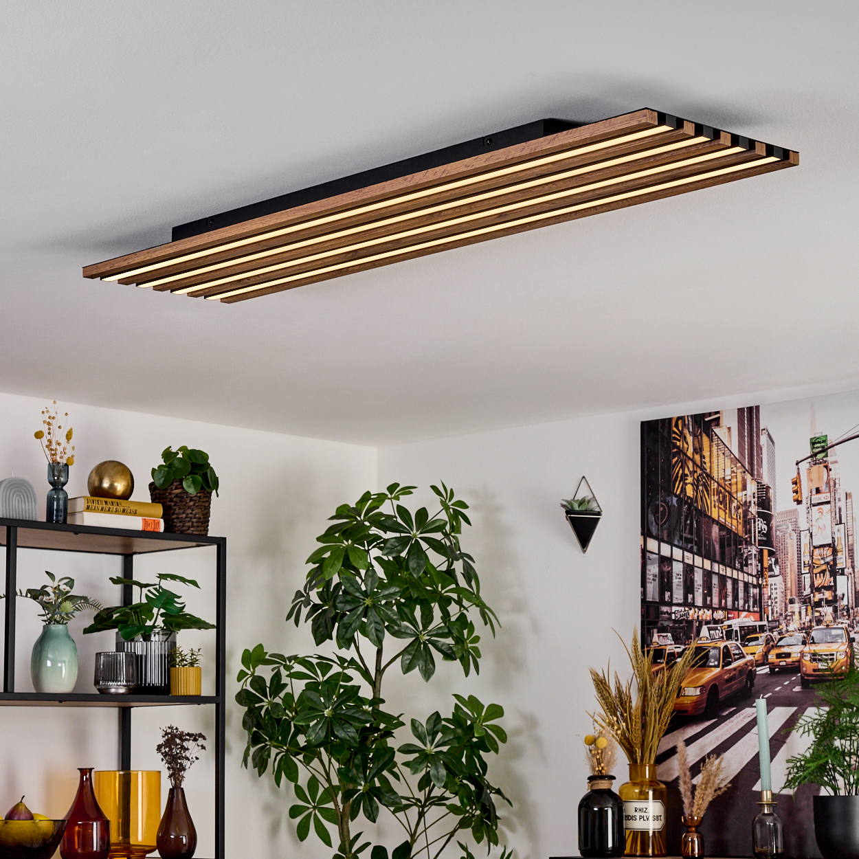 Eine moderne Deckenlampe mit Holzlatten. Ein Regal mit Pflanzen und Dekorationen ist sichtbar. Ein Bild von New York ist an der Wand.