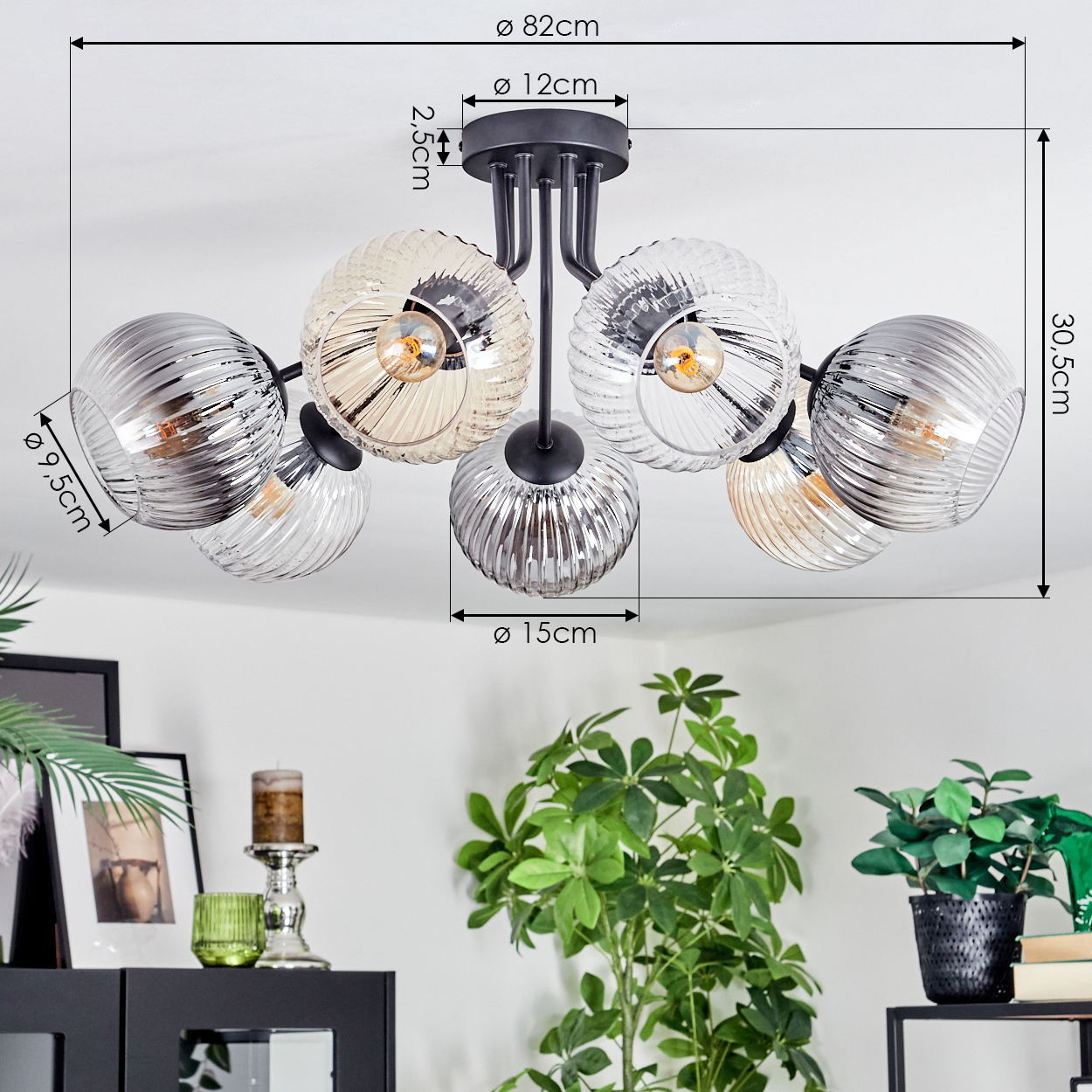 Eine moderne Deckenlampe mit sechs Glasschirmen, silber- und bernsteinfarben. Ein schwarzer Ständer und Armaturen sind ebenfalls sichtbar. Im Hintergrund sind Pflanzen.
