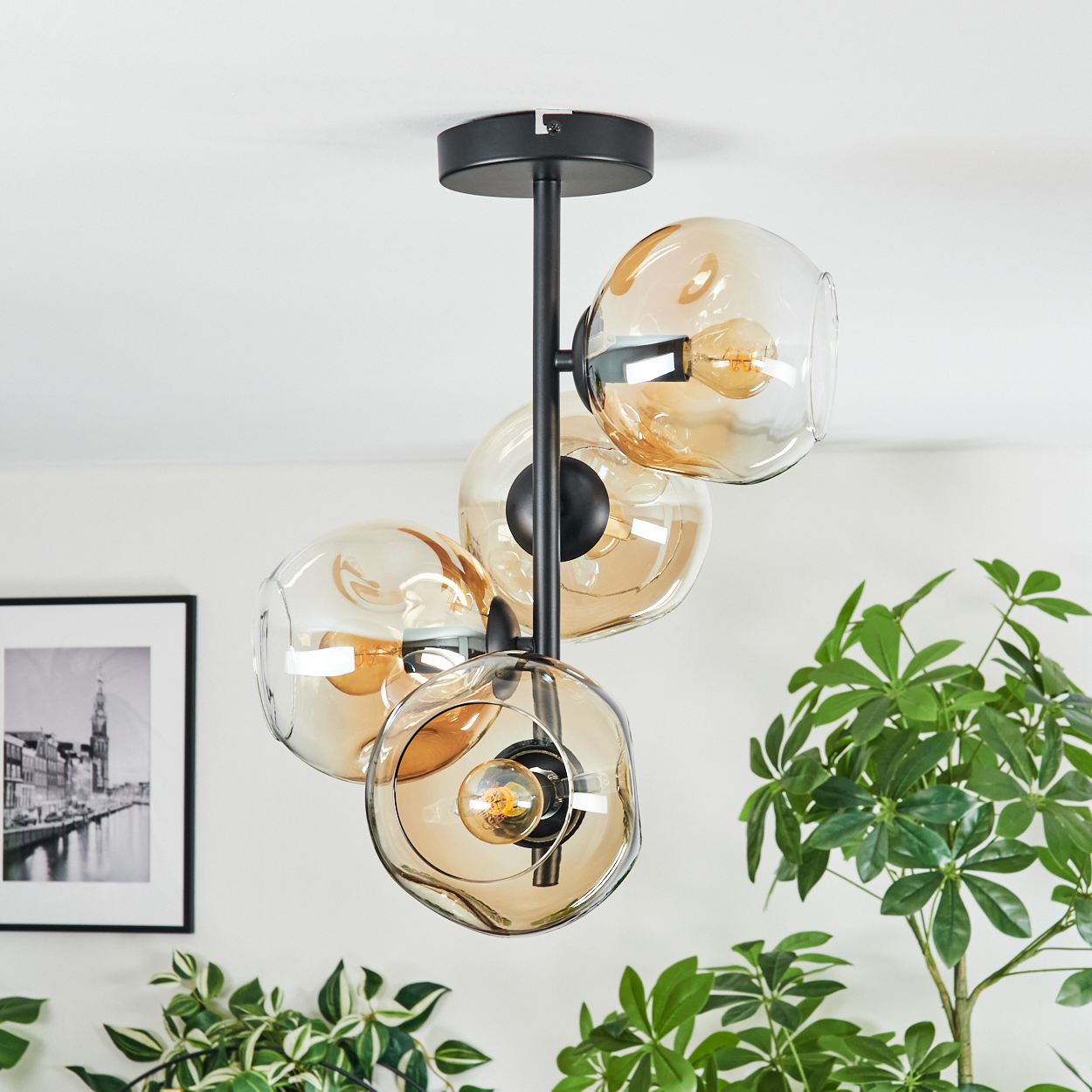 Eine moderne Deckenlampe mit vier bernsteinfarbenen Glaskugeln und einem schwarzen Metallrahmen. Ein schwarz-weiß gerahmtes Bild und Pflanzen im Hintergrund.