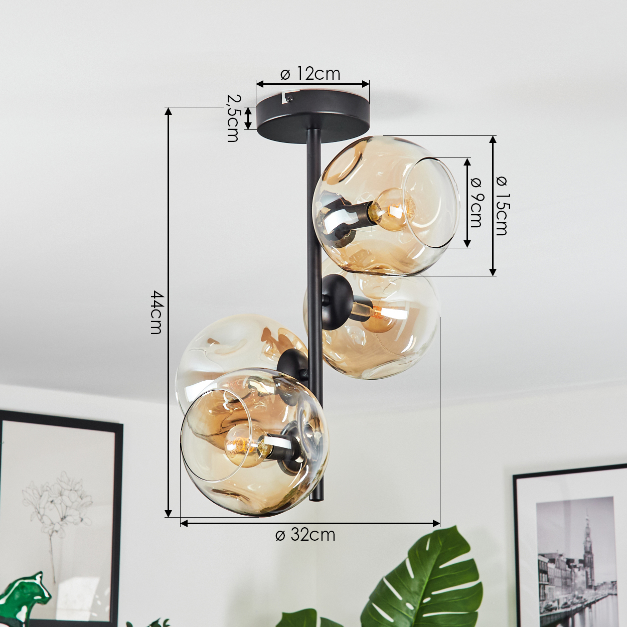 Eine moderne Deckenlampe mit drei bernsteinfarbenen Glaskugeln. Sie hat einen schwarzen Rahmen, Maße sind angezeigt. Hintergrund: Kunst und Pflanzen.