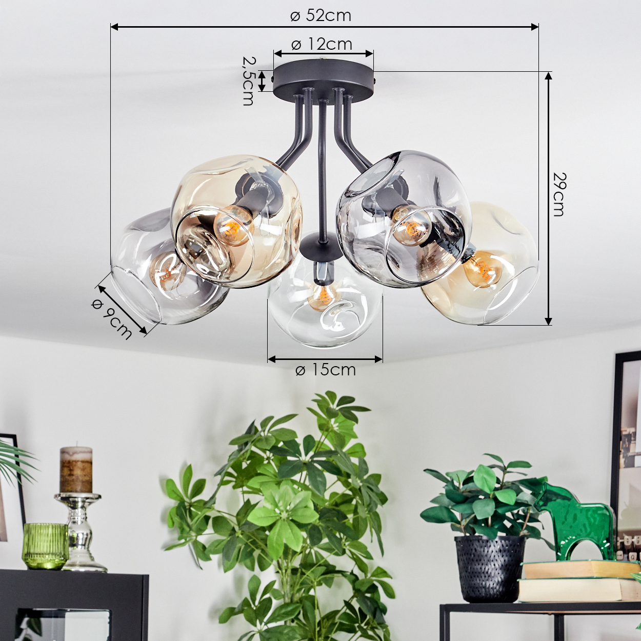 Eine moderne Deckenlampe mit fünf Glaskugeln, Maße beschriftet. Unten sind grüne Pflanzen und Dekorationen zu sehen.
