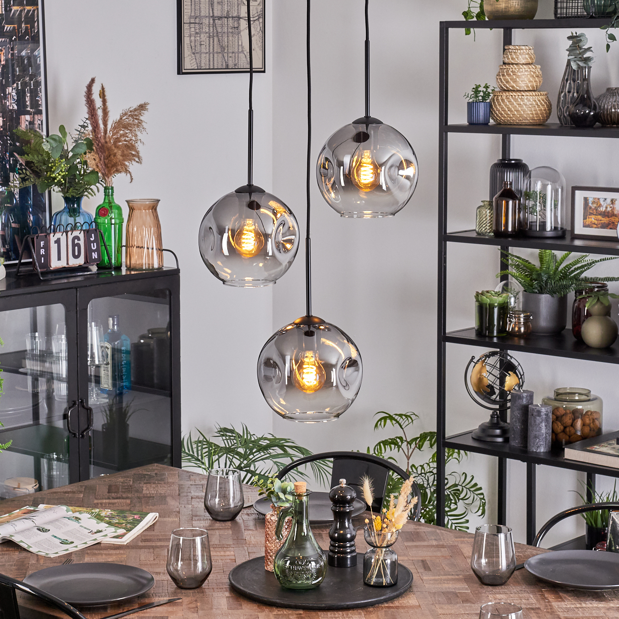 Esszimmer mit Holztisch, drei Hängelampen, schwarzem Regal mit Deko und einem Schrank. Teller, Gläser und Dekoration.