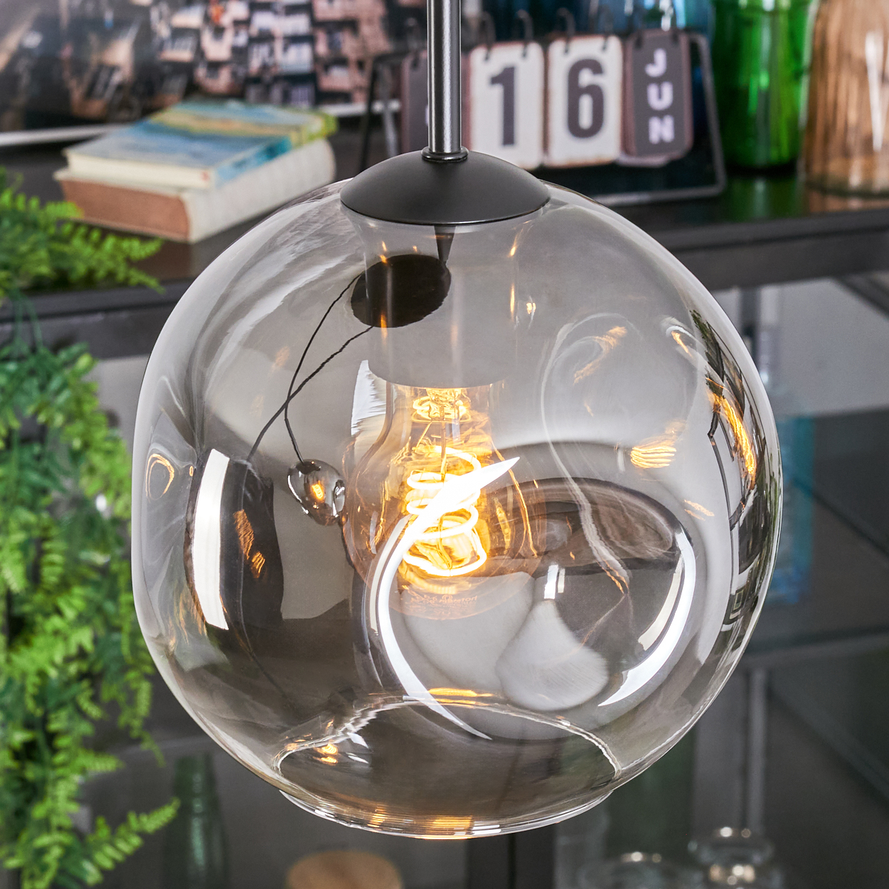 Eine klare, runde Glas-Pendelleuchte mit einer leuchtenden, spiralförmigen Glühbirne im Inneren. Über ein schwarzes Gerät hängt. Der Hintergrund enthält Objekte.