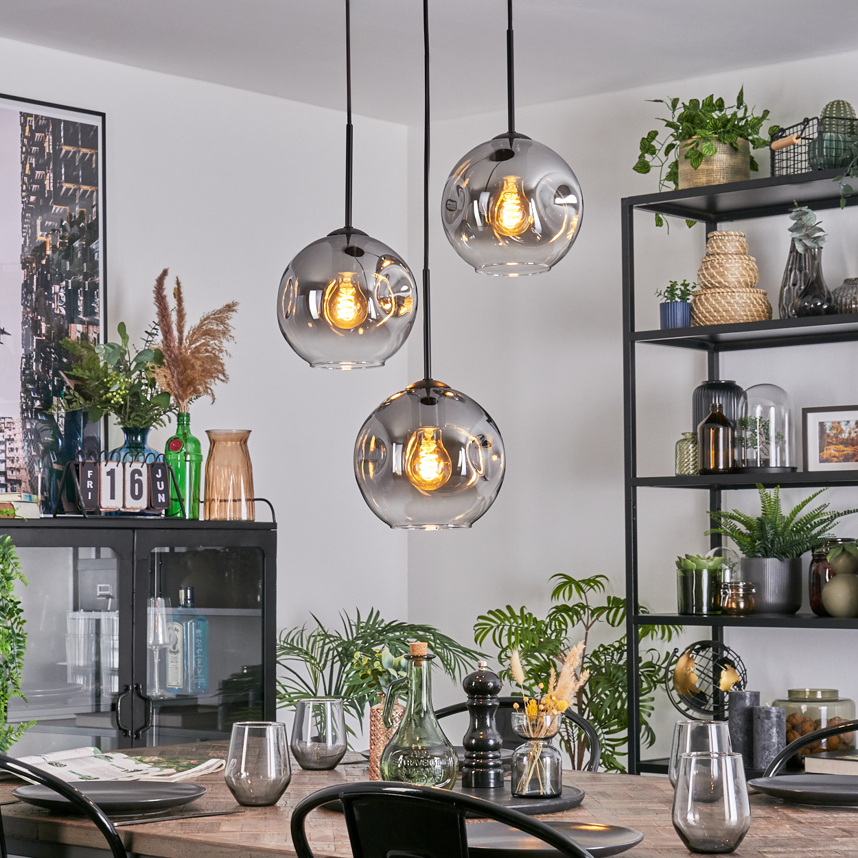 Esszimmer mit drei Hängelampen. Tisch mit Gläsern und Tellern gedeckt. Regale mit Pflanzen und Dekor.