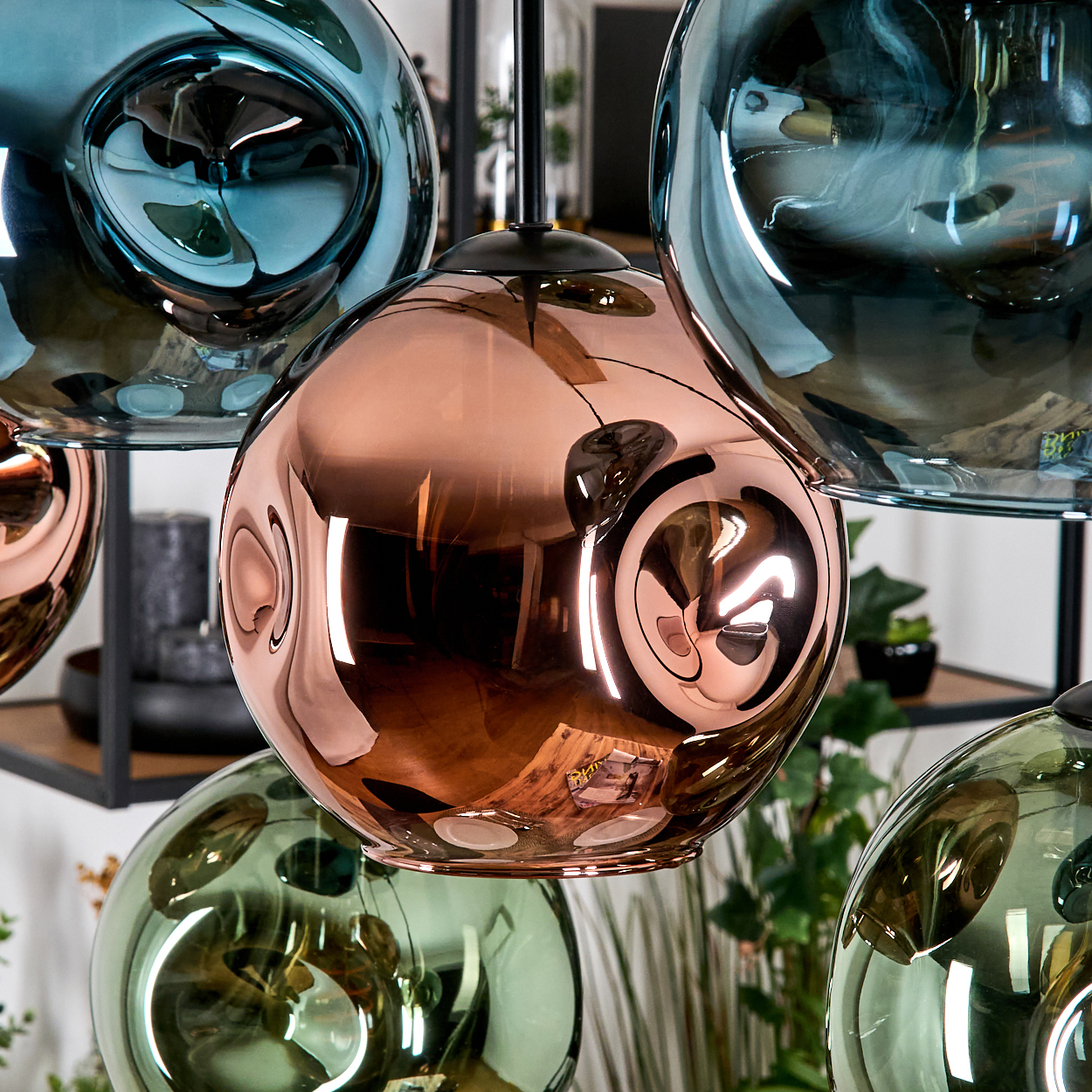 Nahaufnahme von modernen Pendelleuchten. Kupfer- und tealfarbene Glaskugeln reflektieren Licht und Umgebung. Pflanzen im Hintergrund.