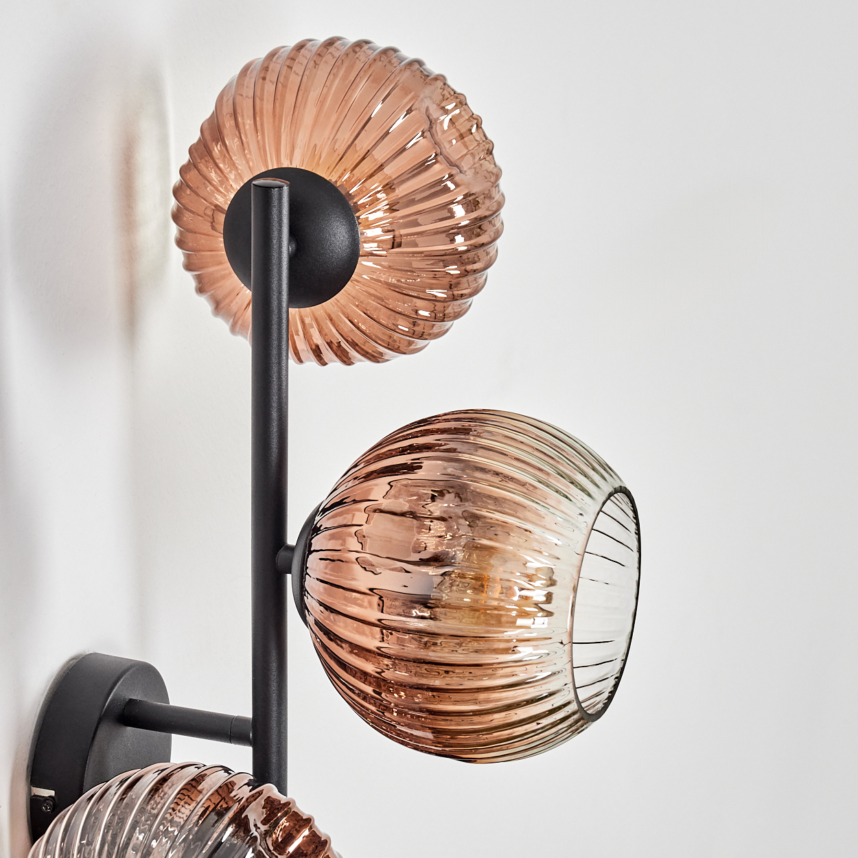 Eine moderne Wandlampe mit drei braunen Glaskugeln. Die Lampe ist auf einem schwarzen Metallrahmen an einer weißen Wand befestigt.
