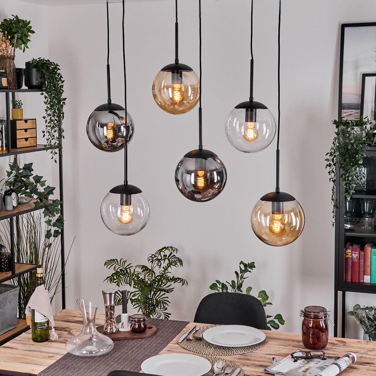 Esszimmer mit sechs hängenden Kugellampen. Tisch mit Tellern, Gläsern und einem Glas. Pflanzen und Regale im Hintergrund.