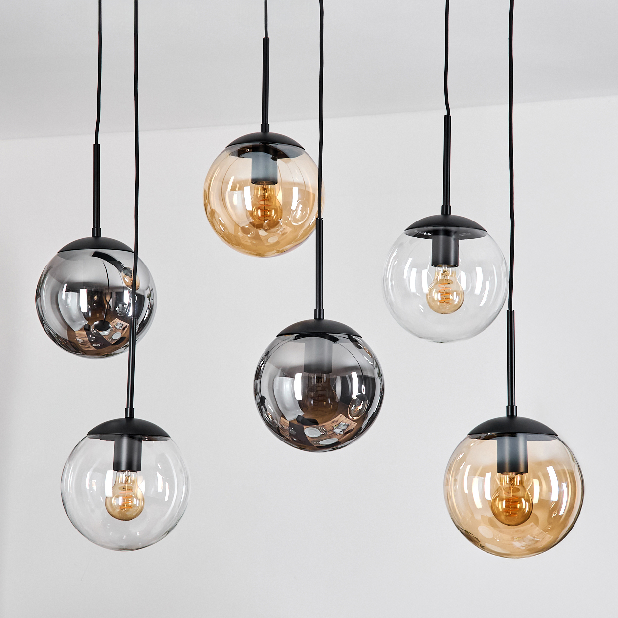 Sechs moderne Pendelleuchten mit Glaskugeln in verschiedenen Farben hängen von der Decke. Schwarze Stiele und Sockel. Weißer Hintergrund.