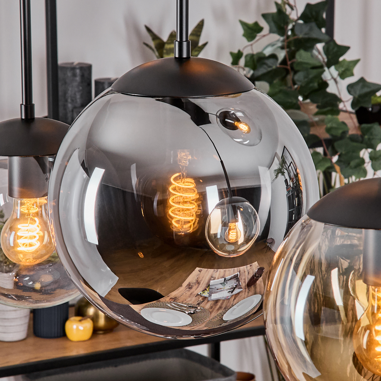 Drei kugelförmige Lampen mit sichtbaren Glühbirnen. Reflektierende Glasschirme. Der Hintergrund zeigt Pflanzen und einen Tisch mit Tellern.