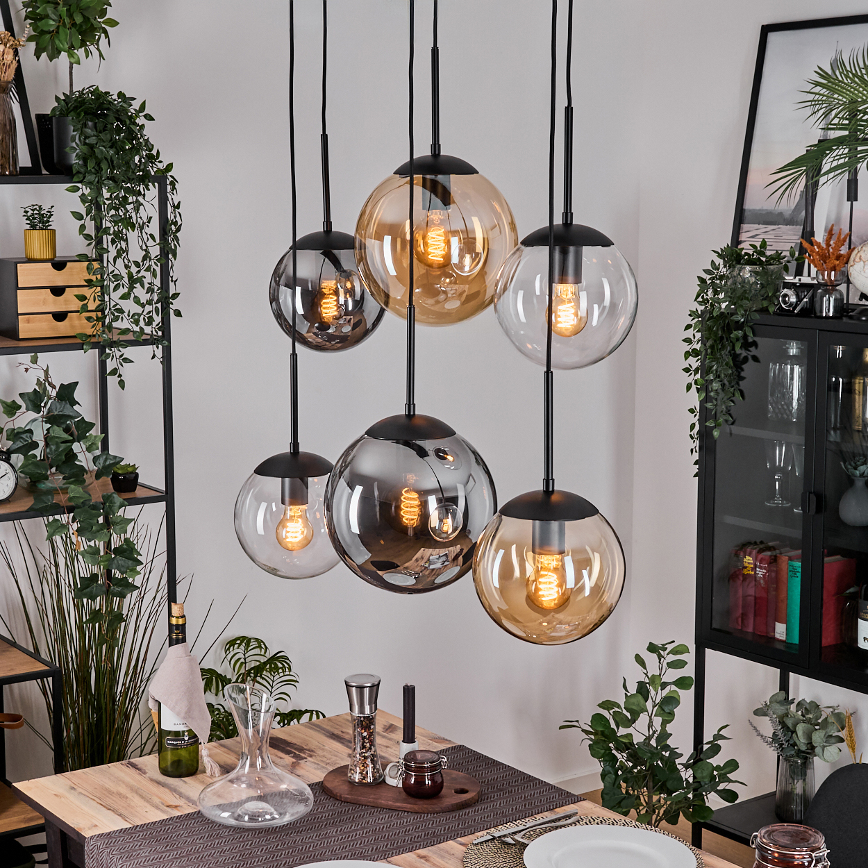 Esszimmer mit Pendelleuchten. Tisch gedeckt mit Karaffe, Pflanzen und Regalen mit Dekor. Kugelleuchten mit Edison-Glühbirnen.