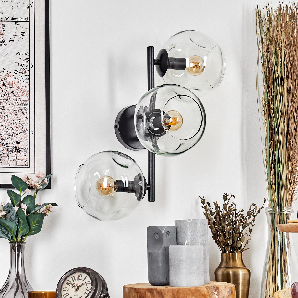 Eine moderne Wandlampe mit drei Glaskugeln. Sie ist an einer weißen Wand mit Dekorationen wie Kerzen und einer Uhr befestigt.