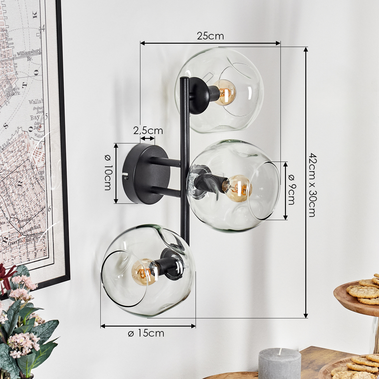 Eine moderne Wandlampe mit drei Glaskugeln, schwarzem Rahmen und sichtbaren Glühbirnen. Sie ist an einer weißen Wand montiert, mit angegebenen Abmessungen.