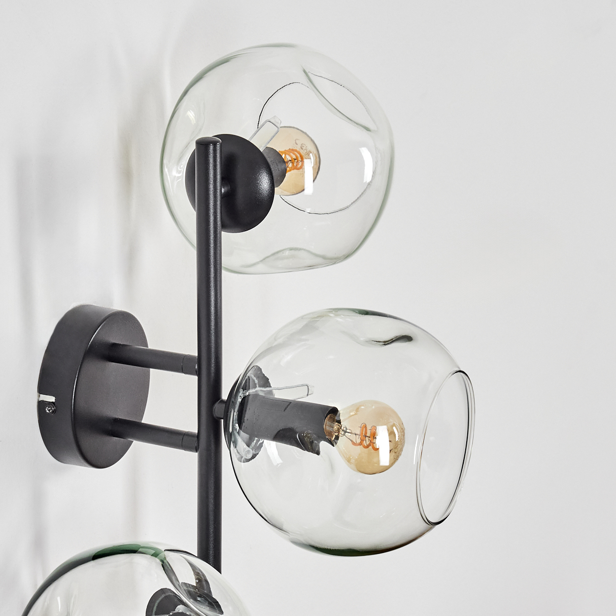 Eine moderne Wandlampe mit drei Glaskugeln und sichtbaren Glühbirnen, montiert auf einem schwarzen Metallrahmen an einer weißen Wand.