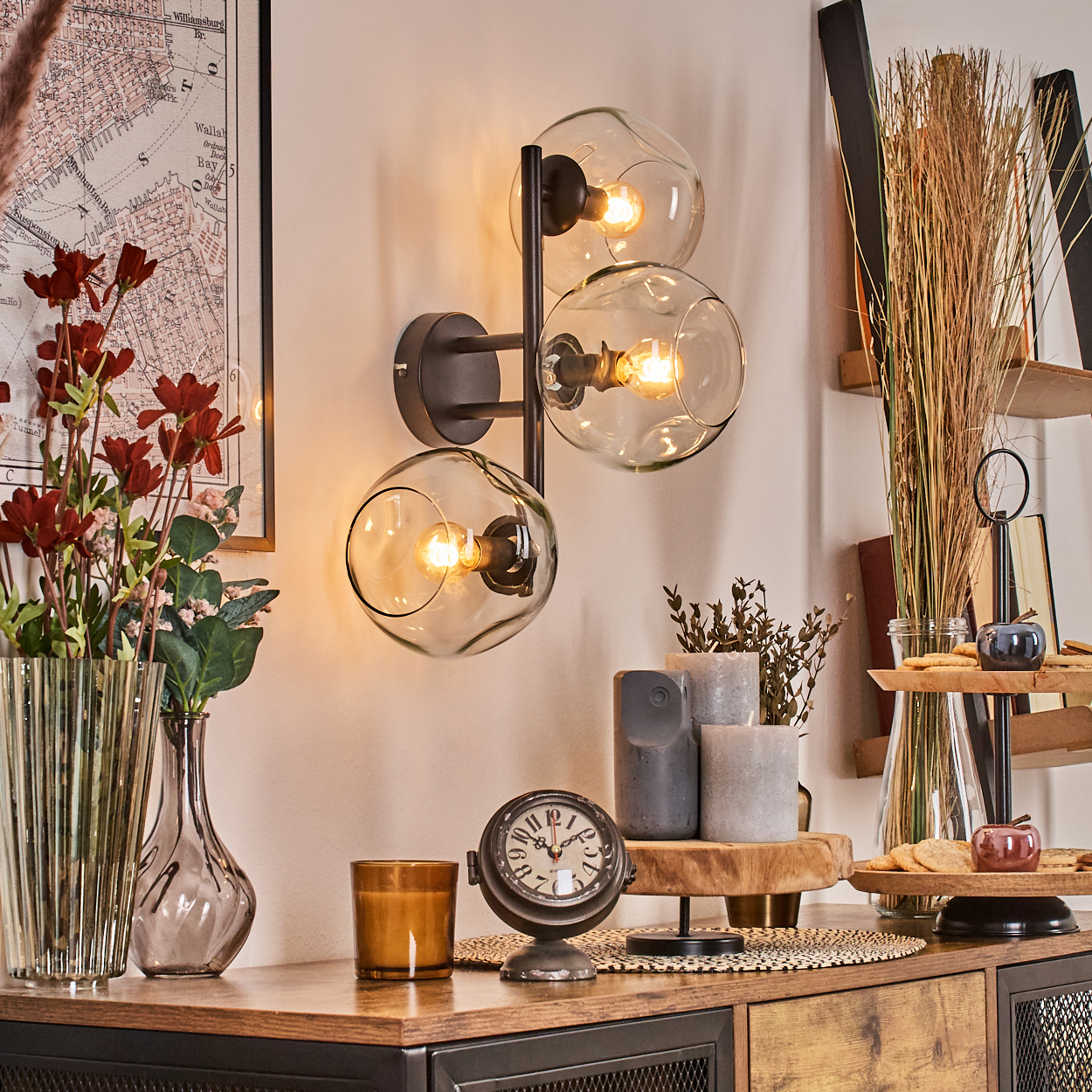 Eine dekorative Wandlampe mit drei Glaskugeln. Auf einem Tisch befinden sich eine Uhr, Kerzen und Vasen mit Blumen. An der Wand hängt eine Karte.
