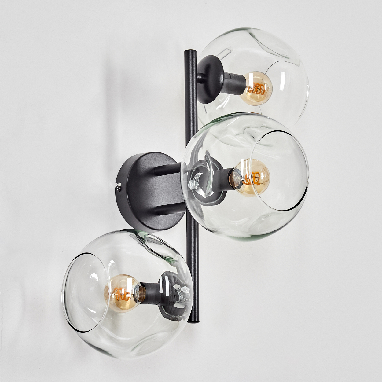 Eine moderne Wandlampe mit drei Glaskugelschirmen, schwarzen Metallarmen und sichtbaren Glühbirnen an einer weißen Wand.