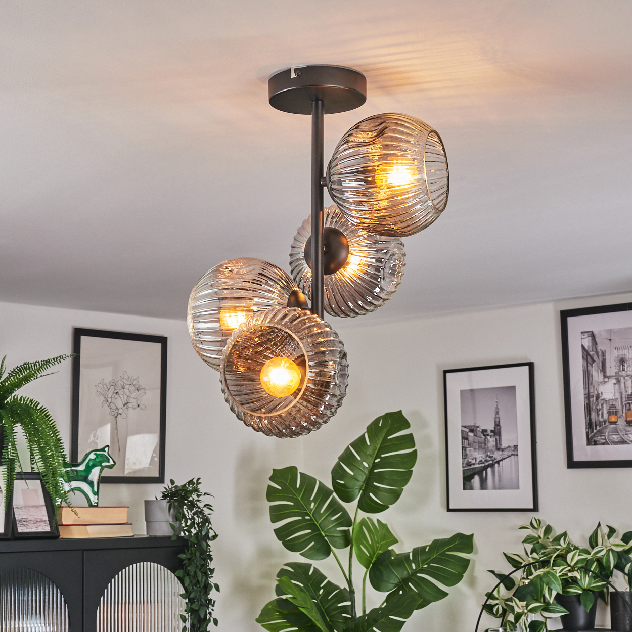 Eine moderne Deckenleuchte mit vier Glasschirmen hängt von der Decke. Im Hintergrund ein Raum mit Bildern und Pflanzen.