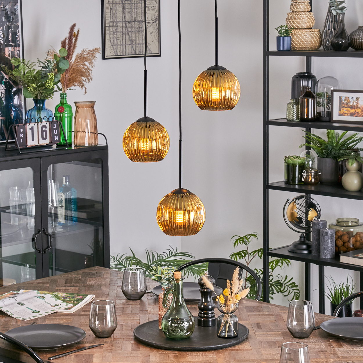 Esstisch mit goldenen Pendelleuchten. Tisch mit Tellern, Gläsern und Dekor. Regale im Hintergrund mit Pflanzen und Dekor.