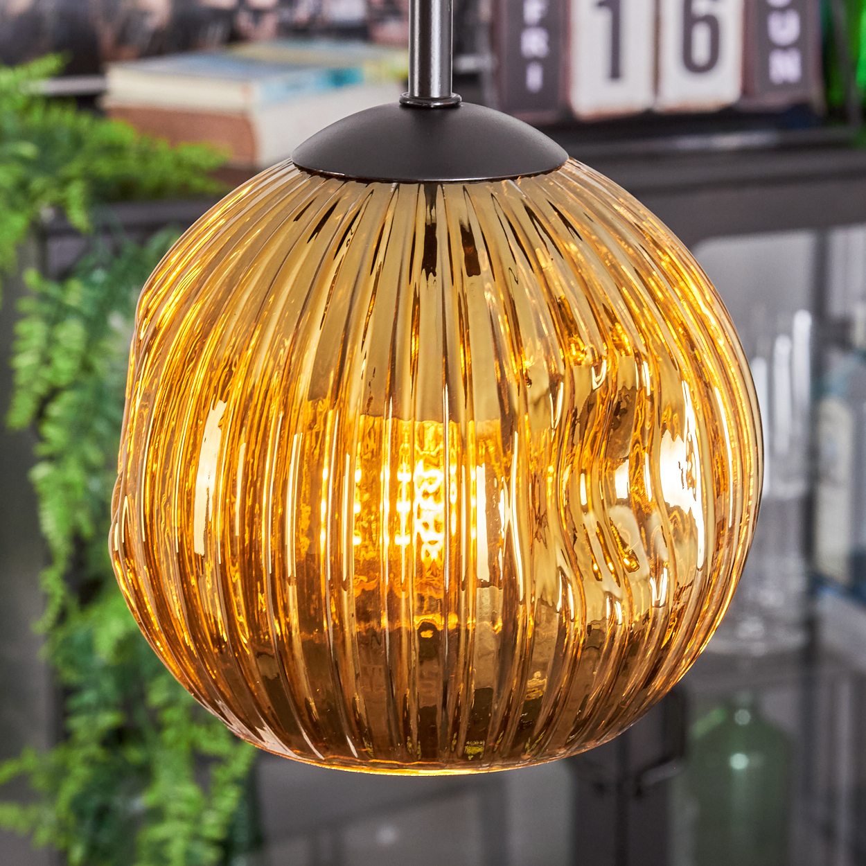 Eine goldene, gerippte Glas-Hängeleuchte hängt von der Decke. Grüne Pflanzen und ein Kalender sind im verschwommenen Hintergrund.