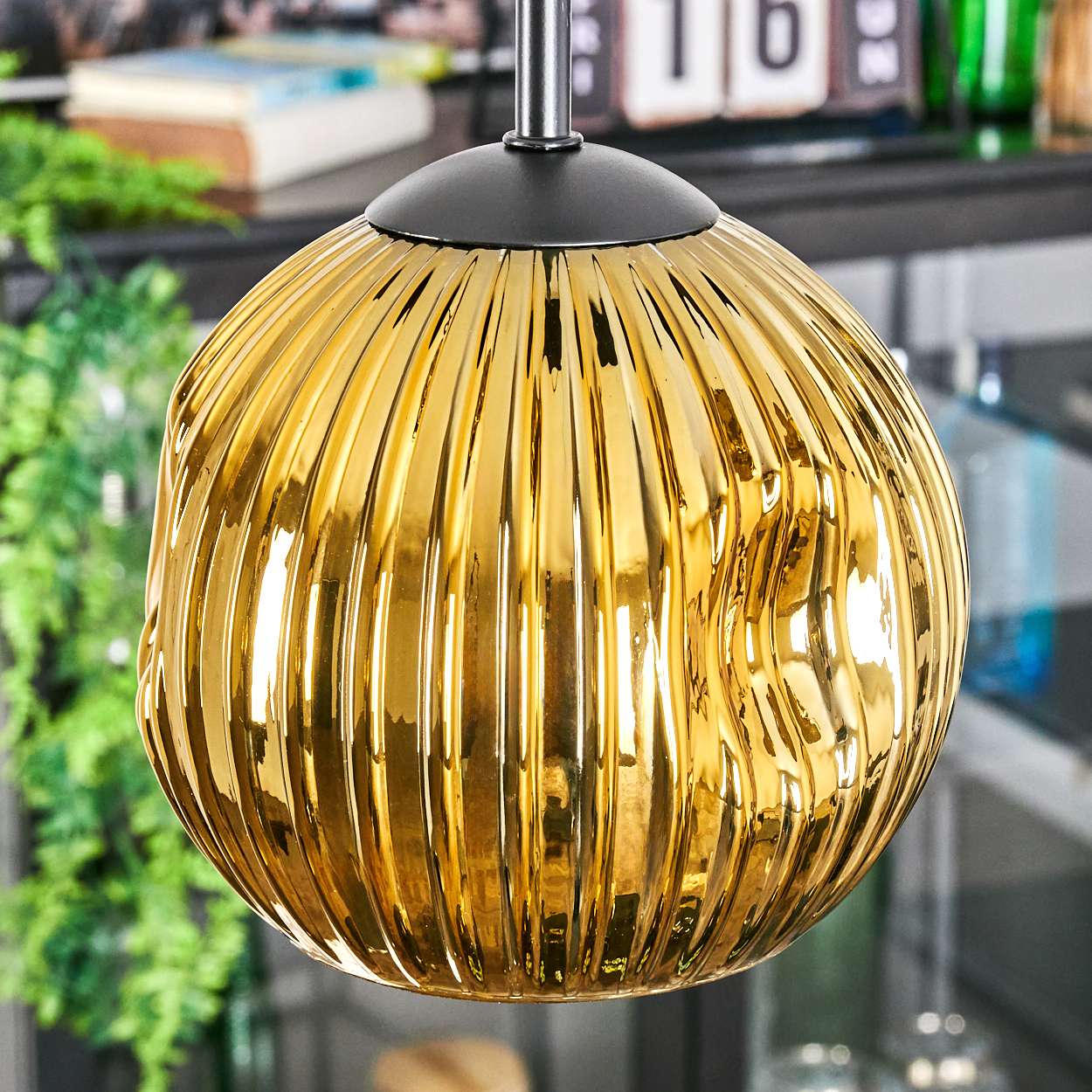 Eine goldene, gerippte Glas-Hängelampe hängt. Der Hintergrund zeigt verschwommenes Grün, ein Regal mit Gegenständen und die Zahl 16.