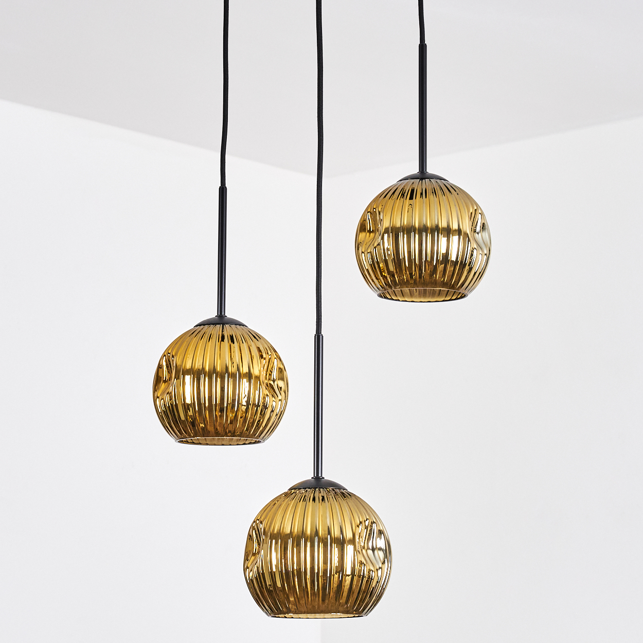 Drei goldene Pendelleuchten hängen an schwarzen Kabeln vor weißem Hintergrund. Die Leuchten haben ein geripptes Design.