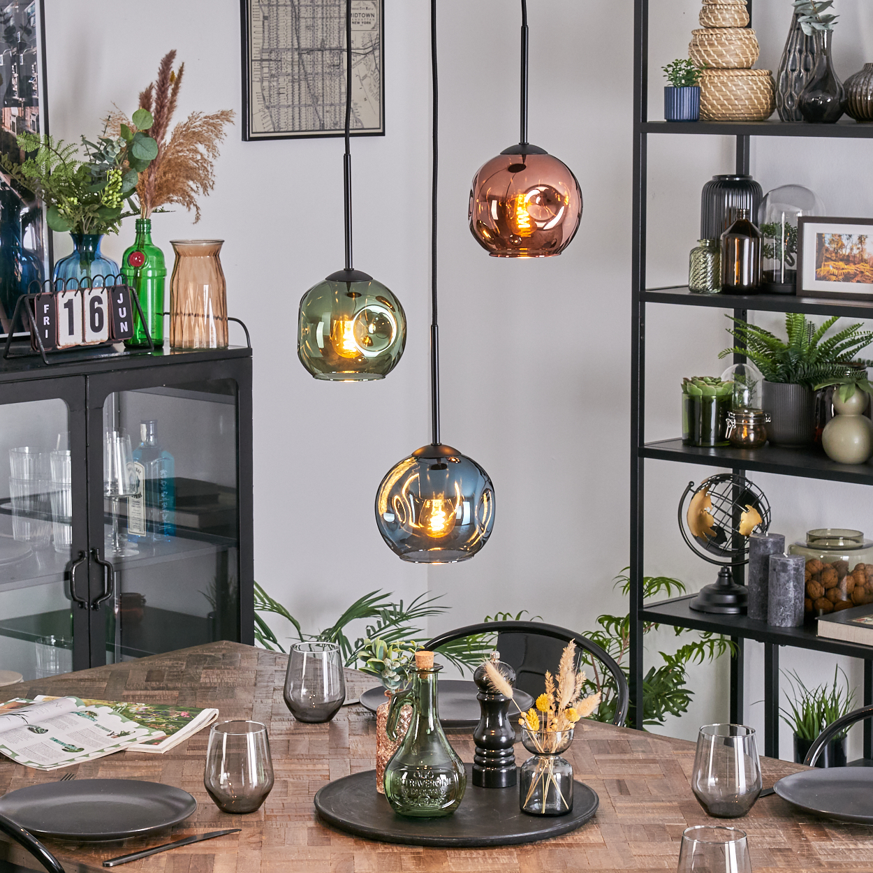 Esszimmer mit Holztisch, Glaslampen und dekorativen Gegenständen. Schwarze Regale mit Pflanzen und Ornamenten im Hintergrund.