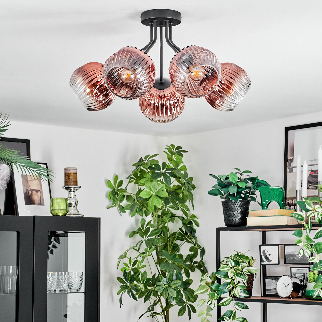 Eine moderne Deckenleuchte mit fünf rosa Glasschirmen hängt über Pflanzen und Dekorationen. Die Szene ist hell und luftig.