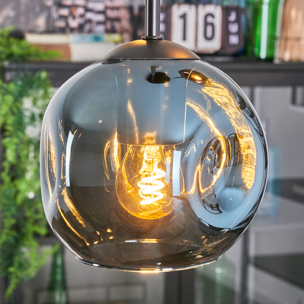 Nahaufnahme einer Glasleuchte mit leuchtender Glühbirne. Das Glas ist rauchgrau und reflektiert die Umgebung. Der Hintergrund hat Objekte.