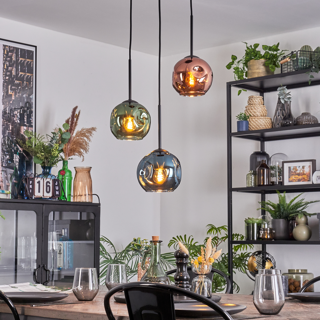 Esszimmer mit drei Pendelleuchten. Tisch mit Gläsern und Tellern. Regale mit Pflanzen und Dekoration im Hintergrund.