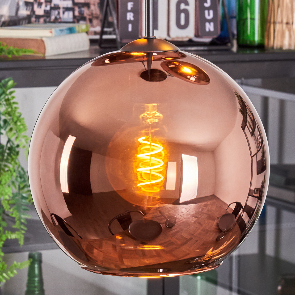 Kupferfarbene kugelförmige Pendelleuchte mit Wendelglühbirne. Das Licht spiegelt den Raum wider. Hintergrund: Kalender und Pflanzen.