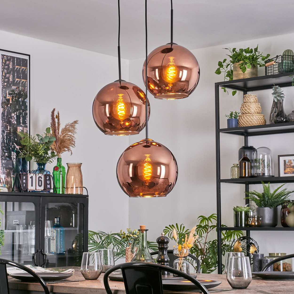 Esszimmer mit kupferfarbenen Kugelleuchten. Tisch mit Gläsern und Tellern gedeckt. Regale mit Pflanzen und Dekoration im Hintergrund.