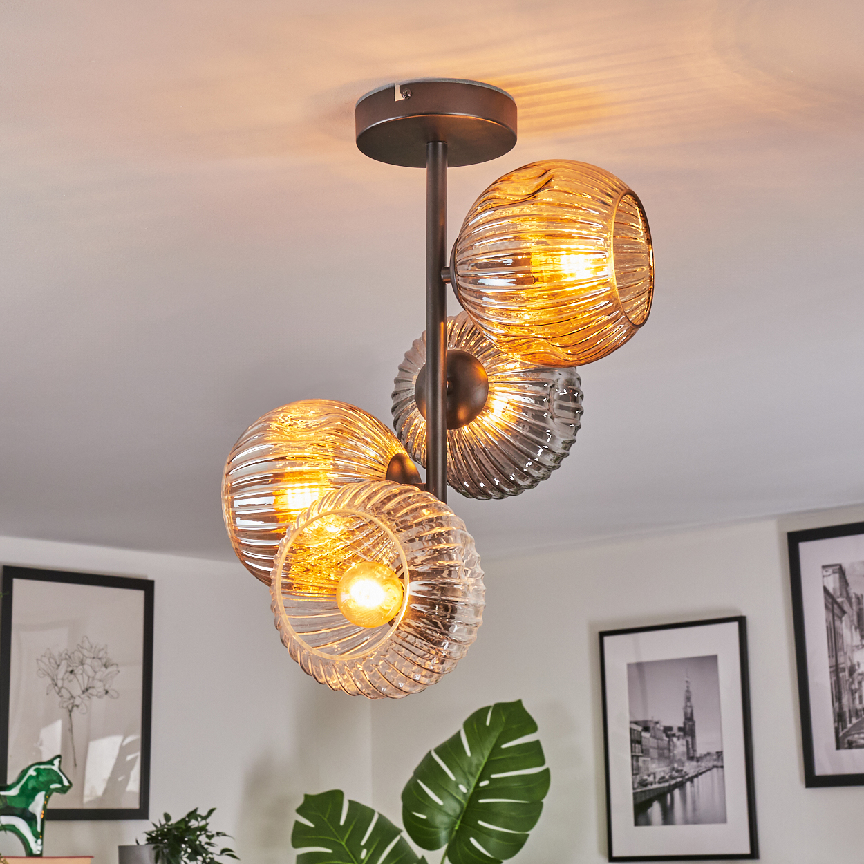 Eine moderne Deckenleuchte mit vier Glaskugelschirmen, die warmes Licht aussenden. Die Leuchte ist an einer weißen Decke angebracht. Kunstwerke an der Wand.