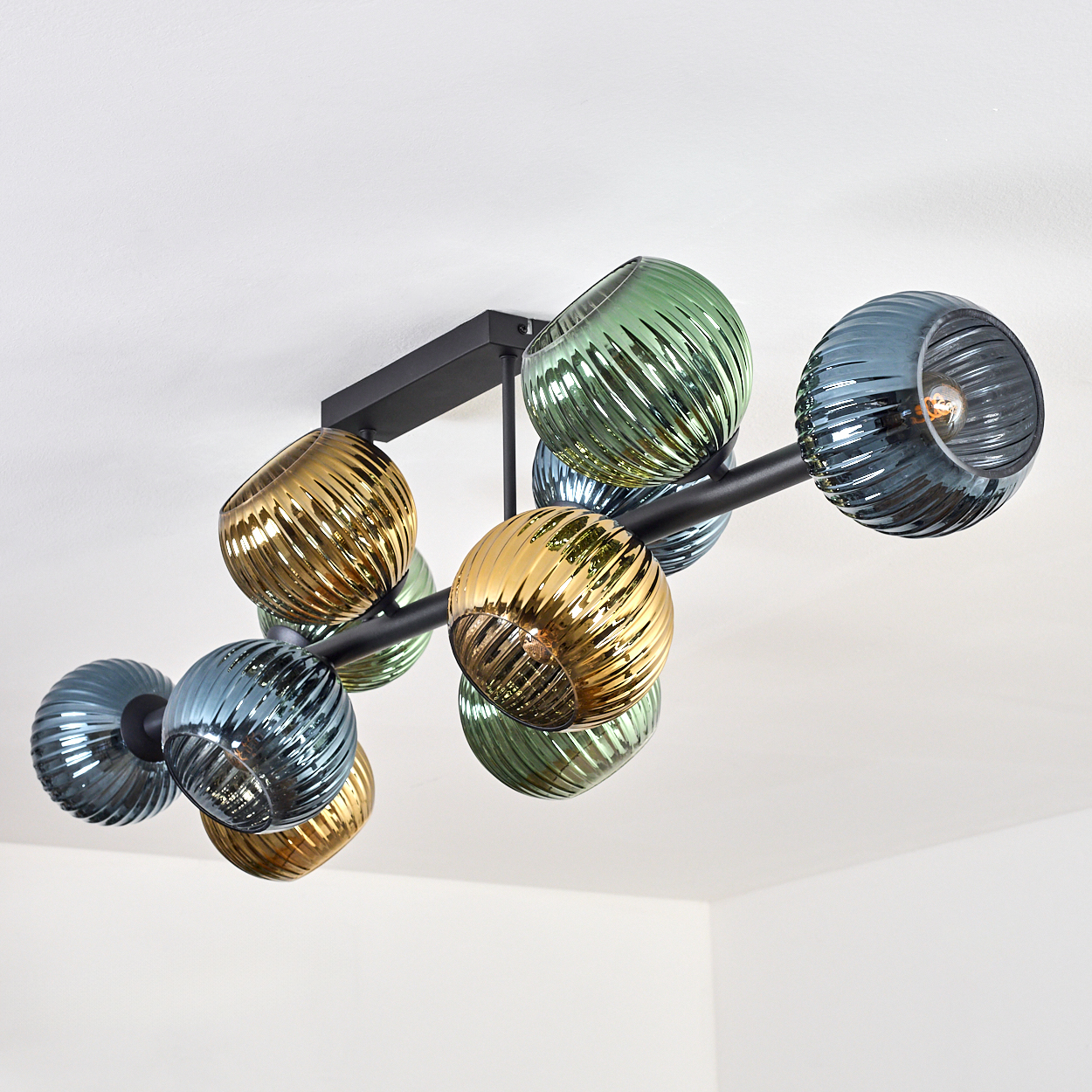 Eine moderne Deckenleuchte mit mehreren geriffelten Glaskugeln in Gold, Grün und Blau. Schwarzer Metallrahmen an einer weißen Decke.